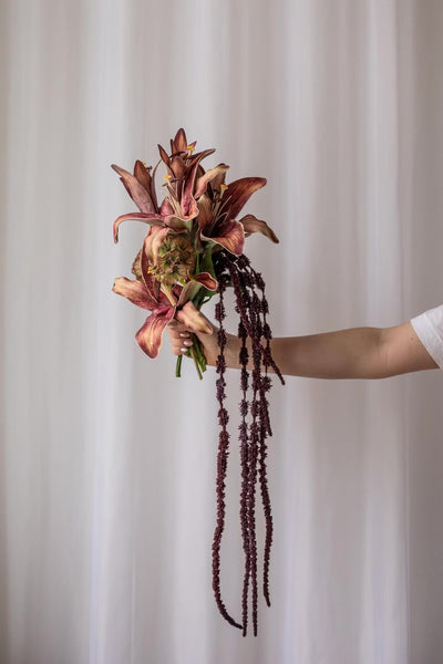 En hånd, der holder en brudebuket med mørkerøde blomster og lange hængende ranker foran en lys baggrund.