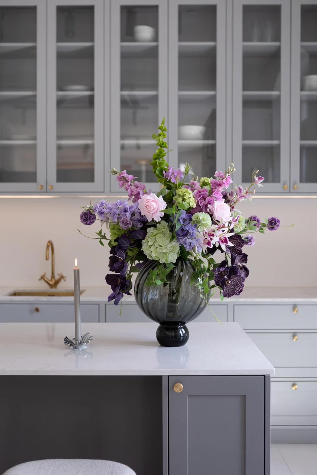 Stor, rund Meadow glasvase i grå med spiralformet mønster. Vasen står på en høj fod. Den er fyldt med en stor buket bestående af lyserøde roser, lilla blomster, grønne hortensiaer og mørkelilla orkidéer. Vasen er placeret på en køkkenø af hvid marmor i et moderne køkken med grå skabe i baggrunden. Til venstre for vasen står et tændt, gråt stearinlys i Lea lysestagen.