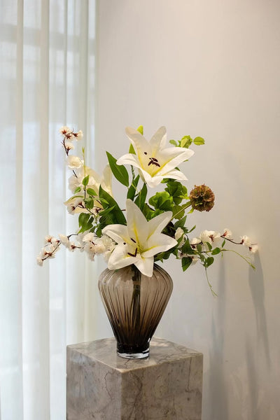 En brun glasvase med en buket af hvide og grønne blomster samt en mørk rødbrun blomst, placeret på en marmorpiedestal foran lyse gardiner.