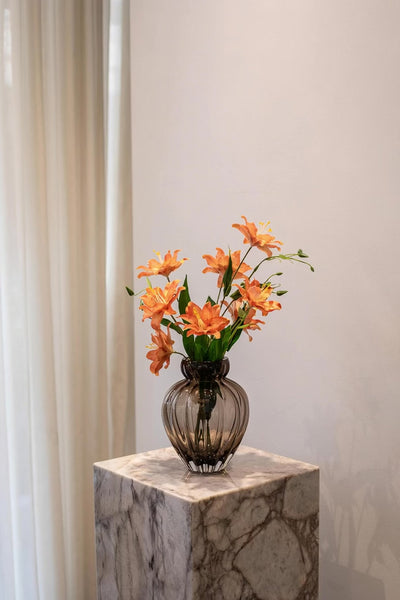 En røgfarvet glasvase med en buket af orange liljer og grønne blade. Vasen står på en marmorpedestal foran et lyst gardin og en lys væg.