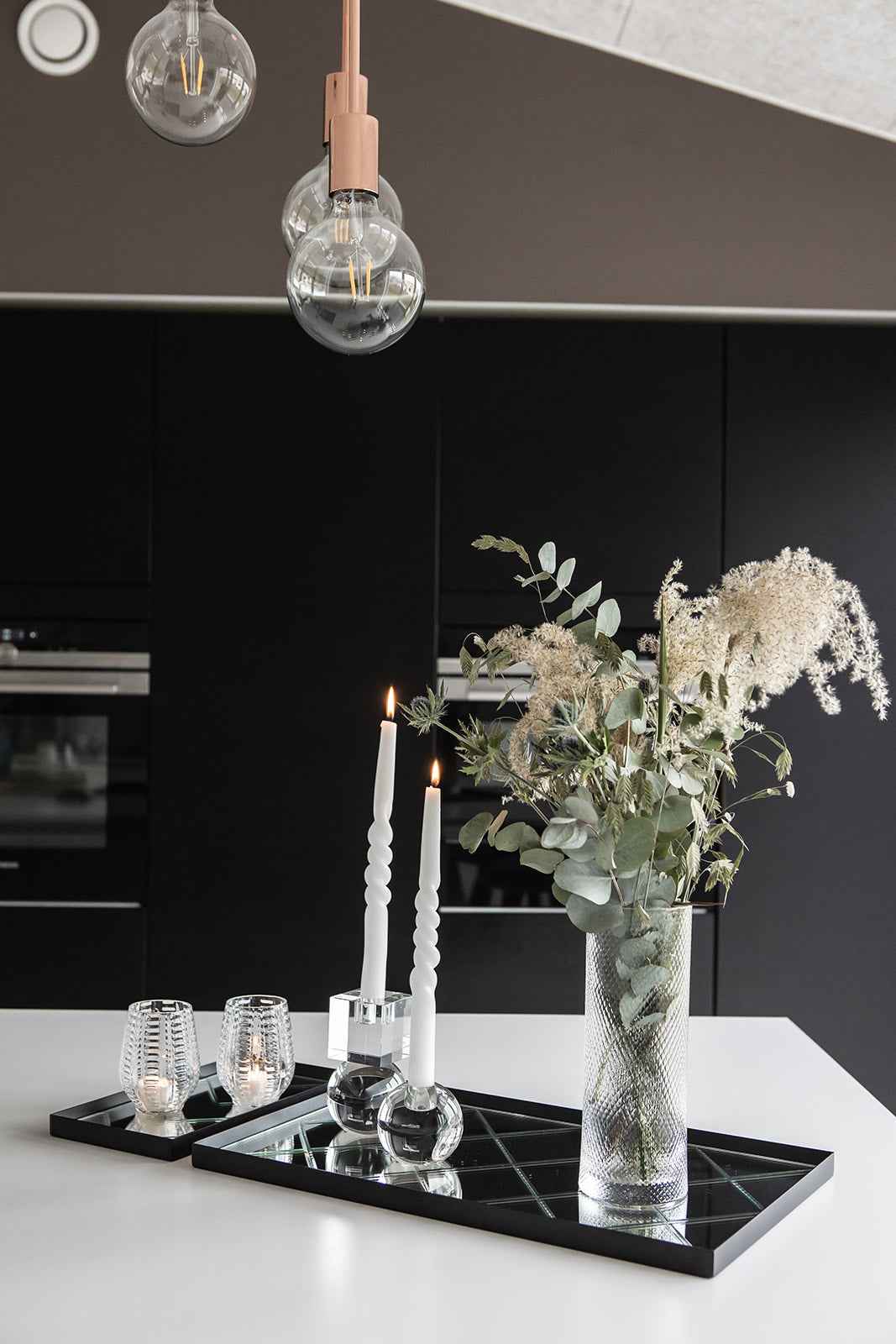 Glasvase med grønt og hvidt blomsterarrangement placeret på en sort spejlbakke. Ved siden af står to tændte snoede hvide lys i glaslysestager med kugleformet fod samt to små fyrfadsstager i rillet glas med tændte lys.