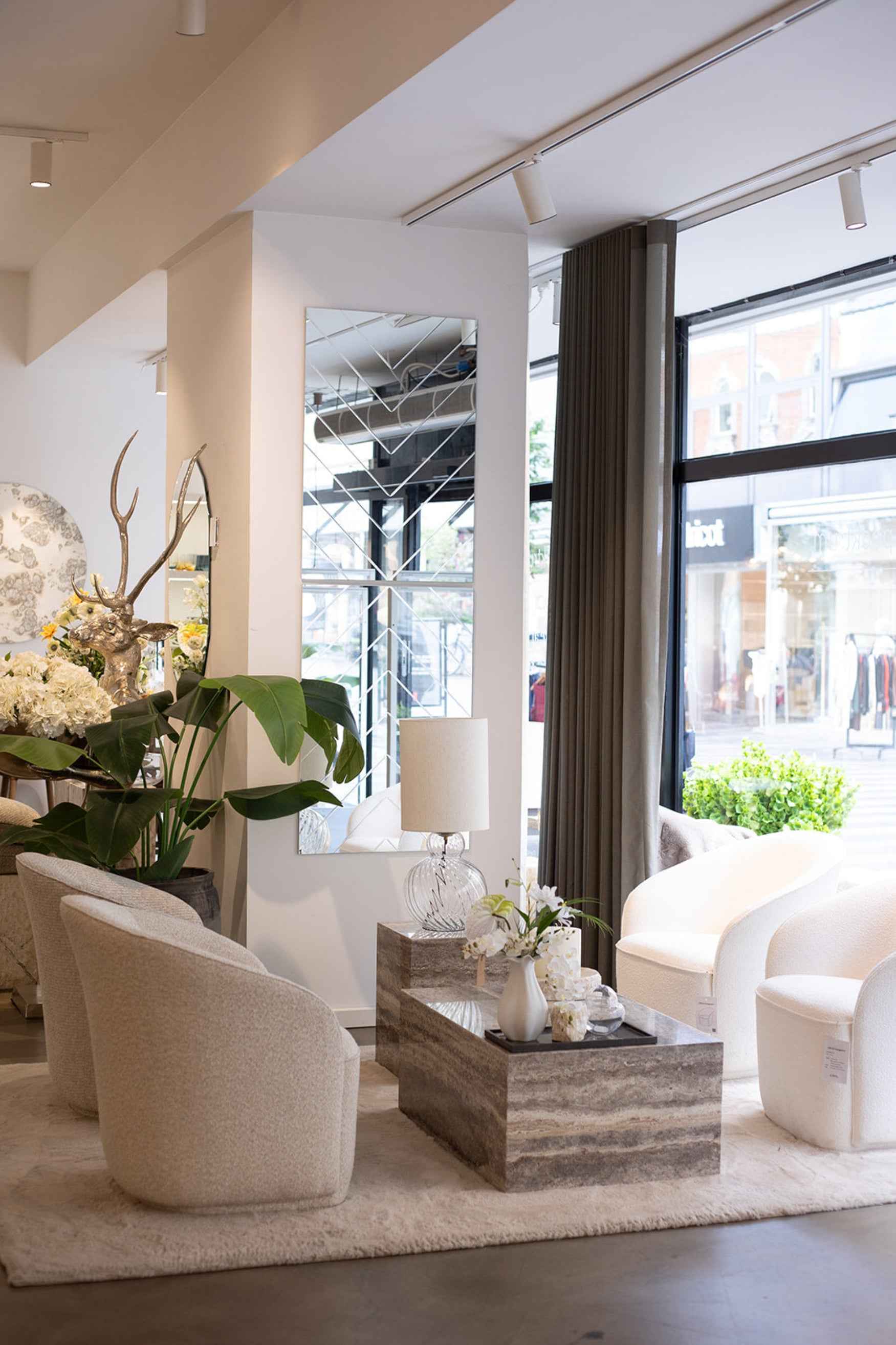 En lys stue med store vinduespartier og moderne indretning. Midt i rummet står to marmorborde, det ene med en bordlampe med klar glasfod og lys stofskærm, det andet med en vase med hvide blomster på en sort bakke. Rundt om bordene står polstrede lyse loungestole på et lyst tæppe. På væggen hænger et rektangulært spejl med mønster, og i baggrunden ses grønne planter og dekorative elementer.