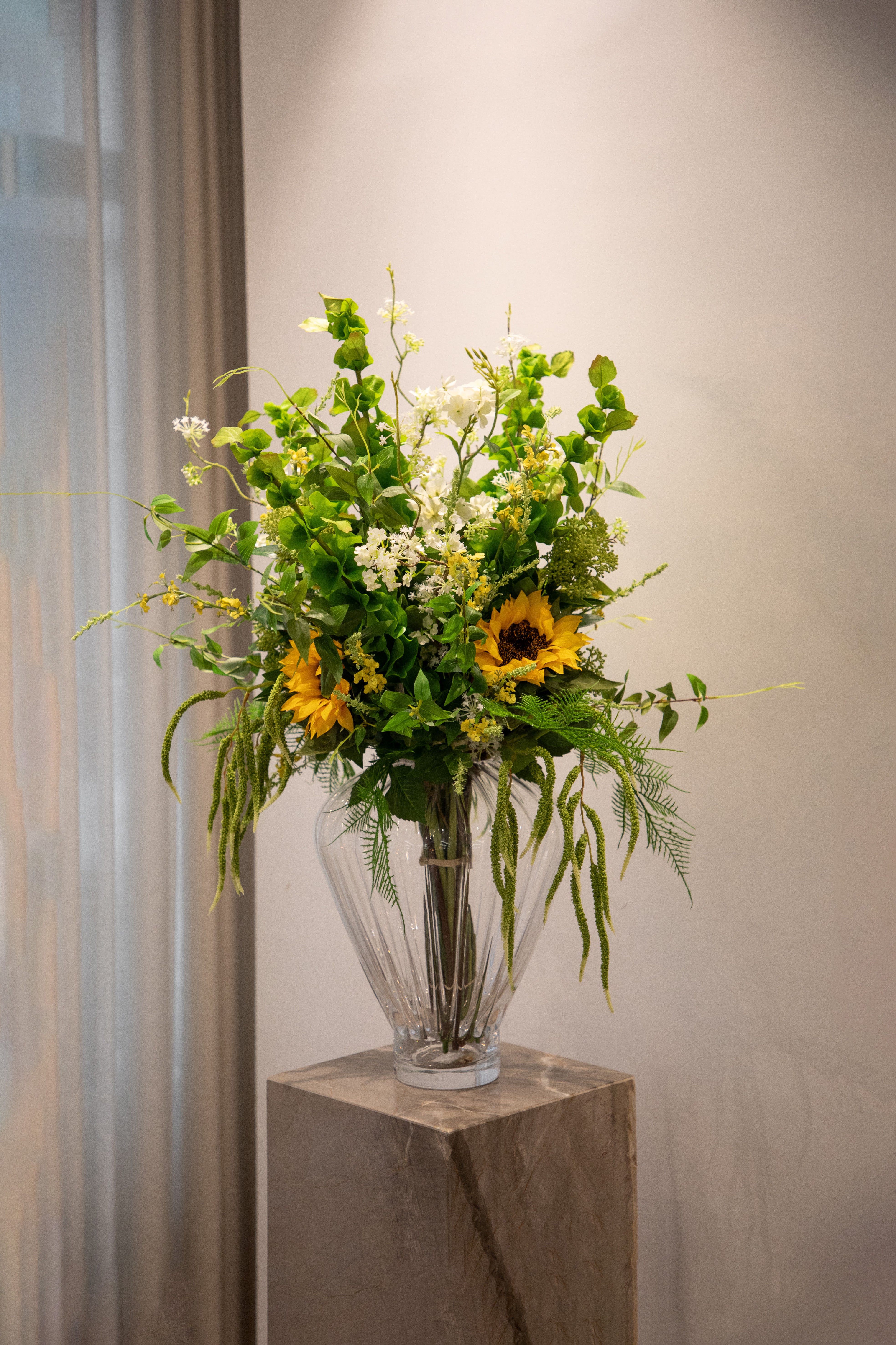 Stor buket af kunstige blomster i grønne, sarte hvide og gule nuancer. Buketten står i en Evelyn gennemsigtig glas vase fra Specktrum ovenpå en marmor pedestal.