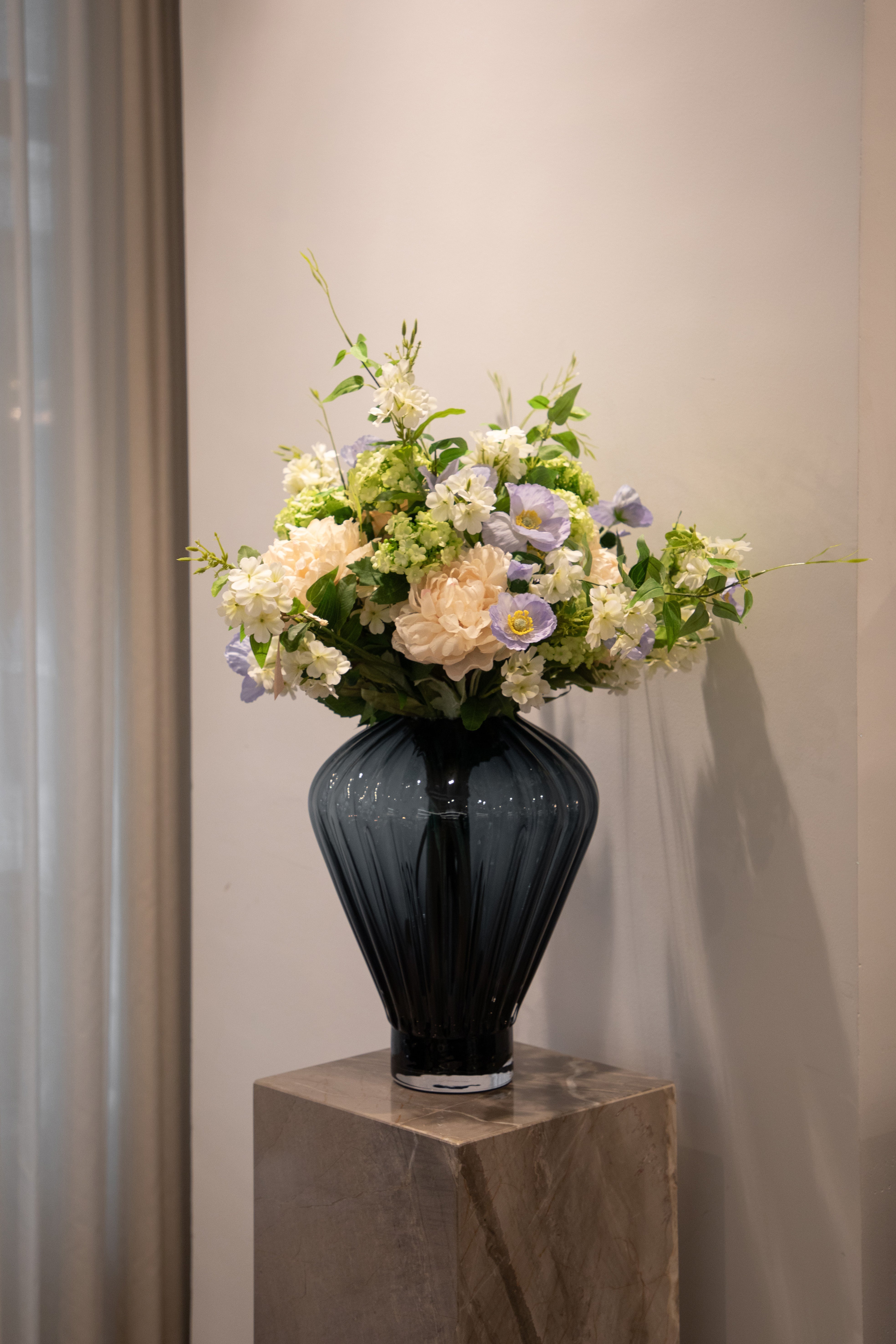 Stor kunstige buket i sarte lilla, hvide, grønne og rosa nuancer. Buketten er placeret i en stor mørkeblå glas vase, der står på en marmor pedestal.