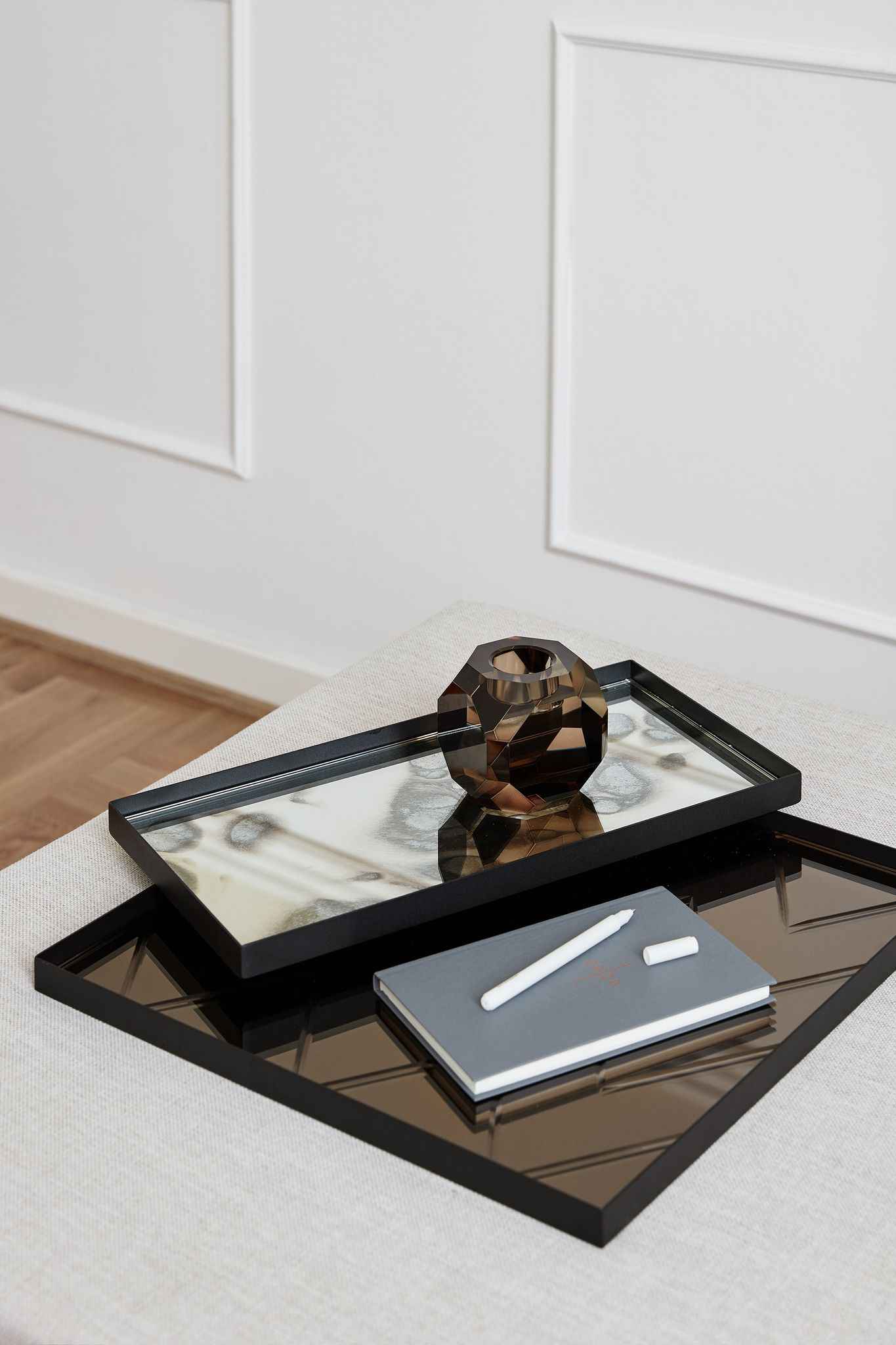 To rektangulære spejlbakker med sort kant placeret oven på hinanden. På den øverste bakke står en brun, facetslebet lysestage i glas. På den nederste bakke ligger en grå notesbog med en hvid pen og kridt ovenpå.