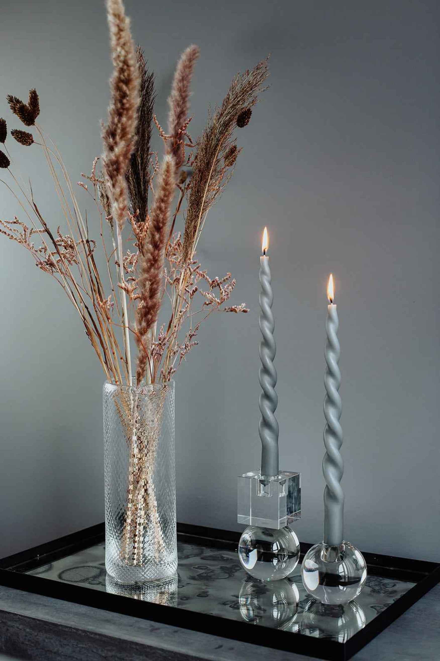 Et dekorativt stilleben med en gennemsigtig glasvase fyldt med tørrede græsser og strå, placeret ved siden af to tændte snoede grå stearinlys i moderne glaslysestager på en sort bakke.