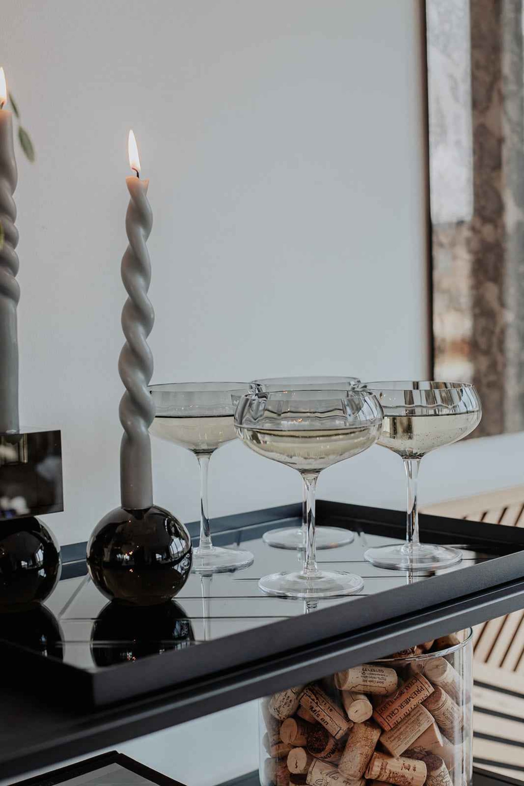 Et dækket serveringsbord med en tændt snoet grå stearinlys i en sort rund lysestage ved siden af fire champagneglas med mousserende vin, ovenpå en bakke med korkpropper i en glascylinder nedenunder.