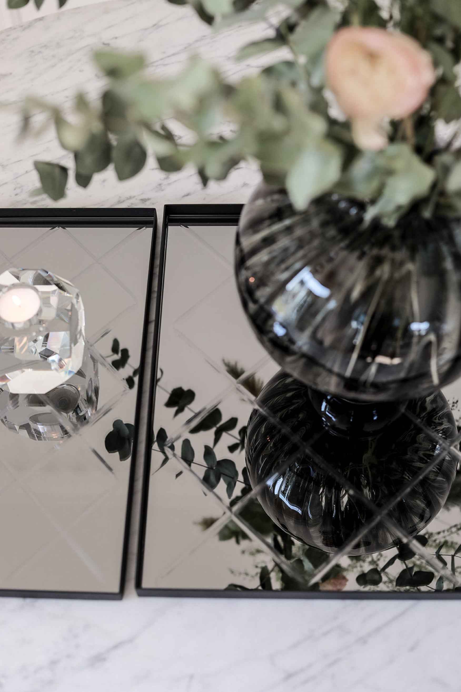 Nærbillede af to sorte rektangulære bakker med spejloverflade, dekoreret med en riflet glasvase med grønne grene og en krystal fyrfadsstage.