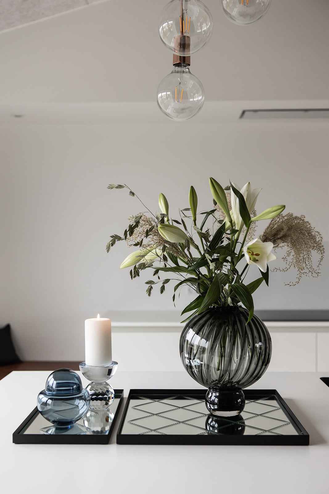 Sort-rillet glasvase med hvide liljer og grønne grene placeret på en kvadratisk spejlbakke. Ved siden af står en klar lysestage med hvid bloklys og en blå glasdekoration på en anden spejlbakke.