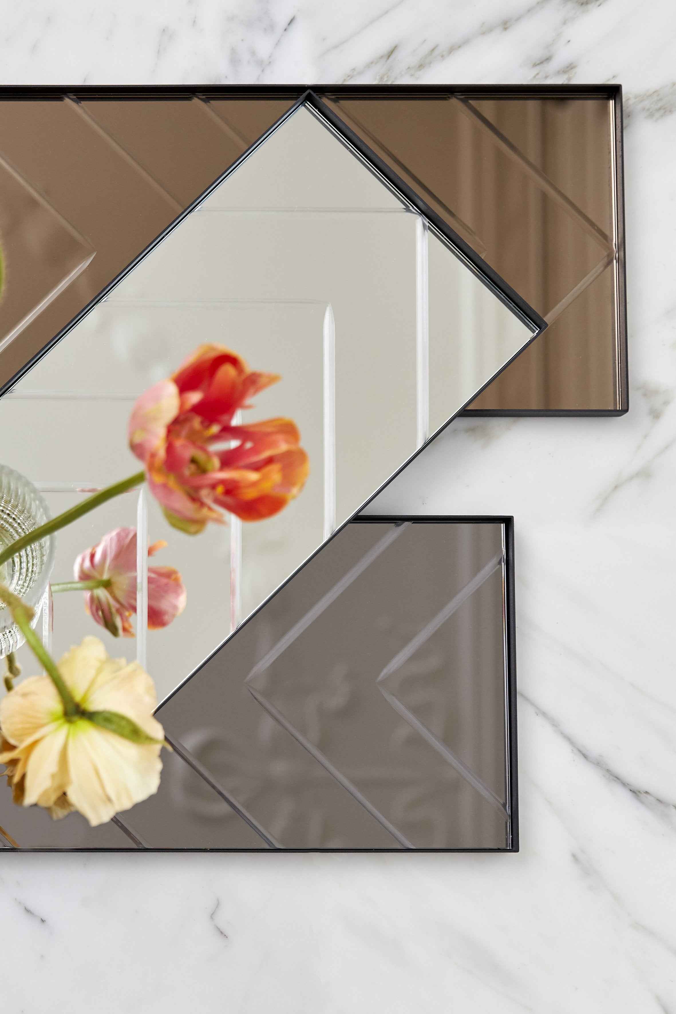 To rektangulære bakker med spejloverflade ligger forskudt oven på hinanden på en marmorbordplade. I spejlingen ses blomster i gule og røde nuancer.