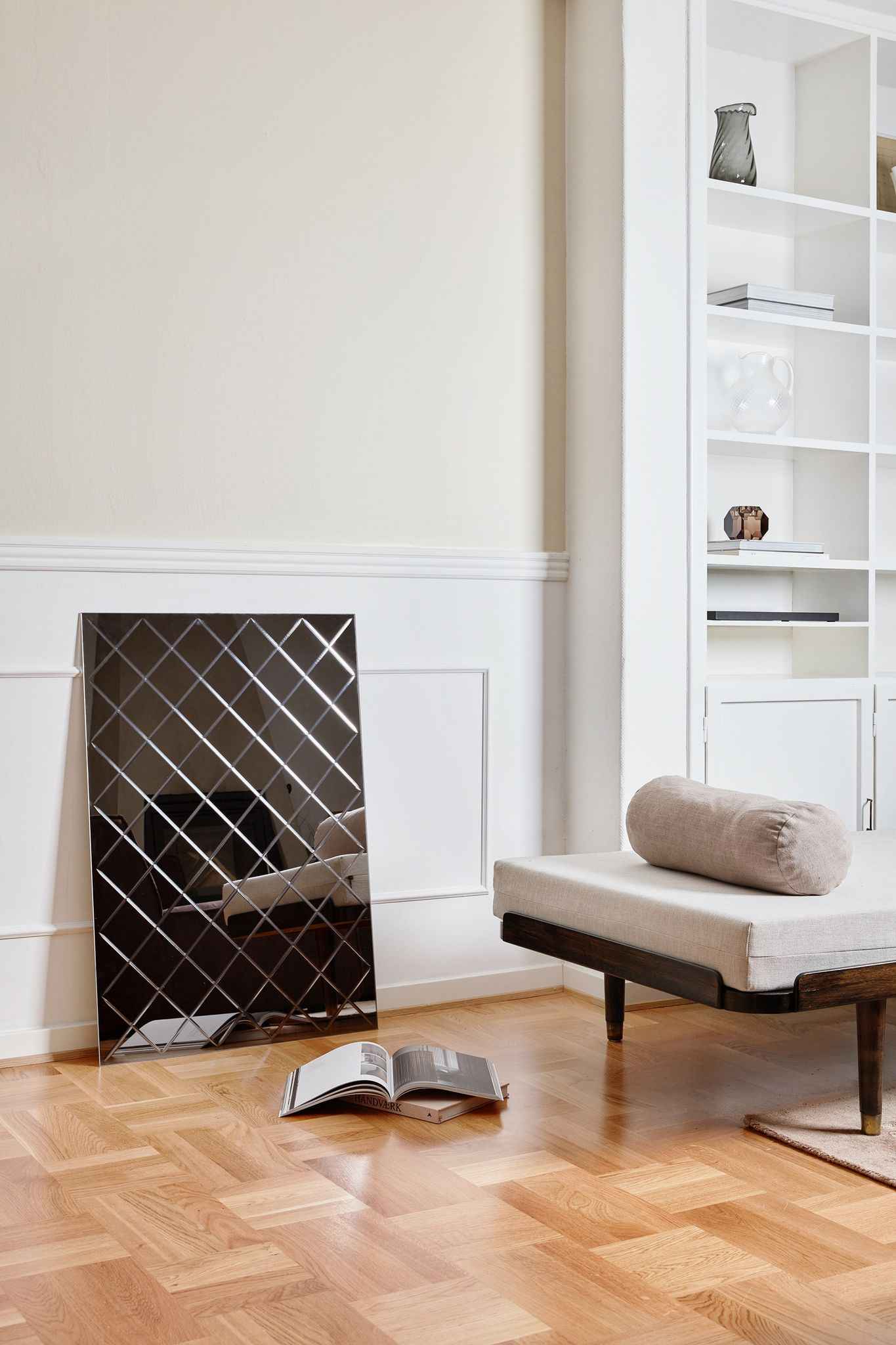 Rektangulært spejl med kvadratisk gittermønster står op ad en væg i et lyst rum. Foran ligger en åben bog på trægulvet, og ved siden af står en bænk med lys hynde og rund pude. I baggrunden ses en hvid reol med dekorative objekter.
