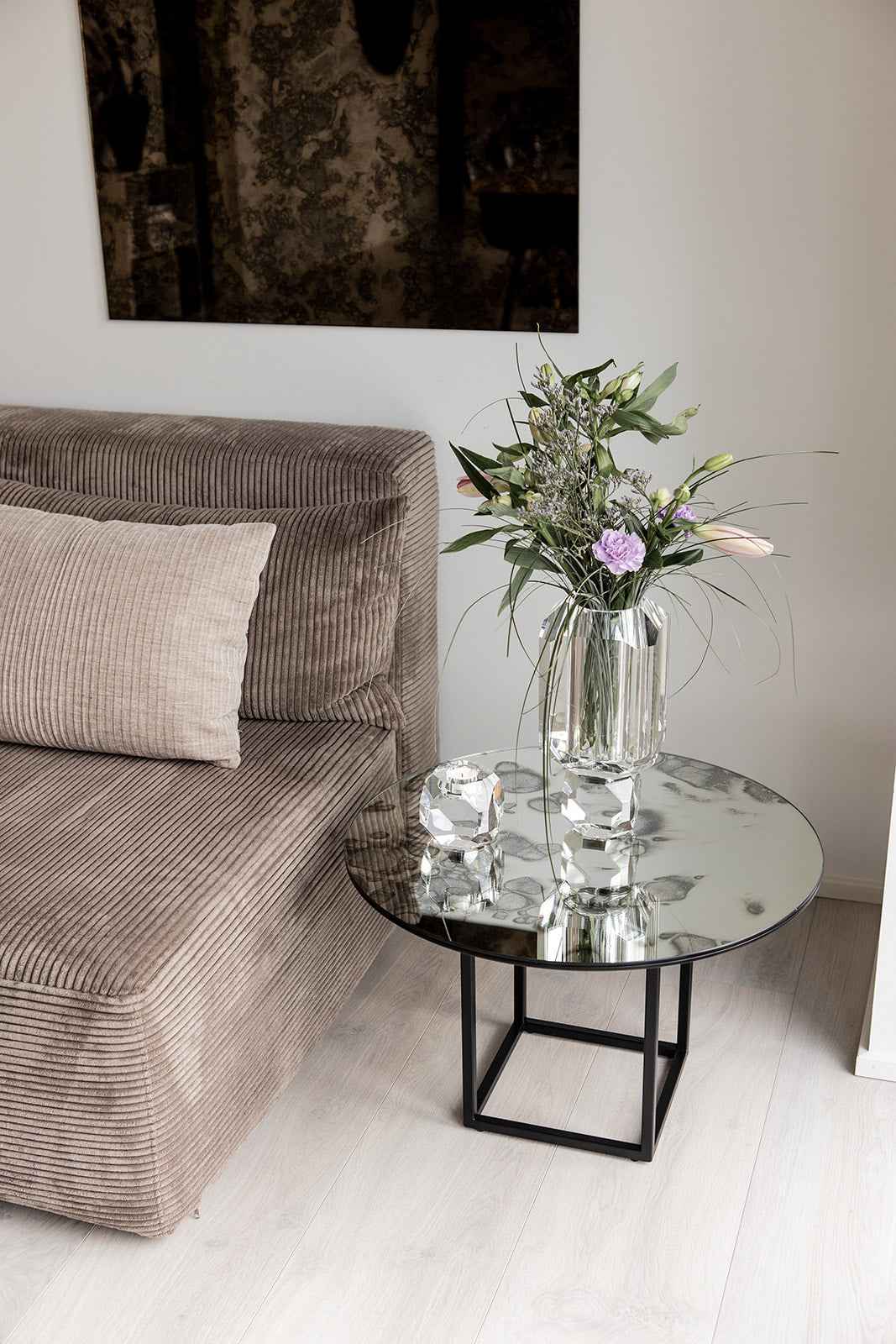 Rundt sofabord med mønstret spejlglasplade og sorte metalben, dekoreret med en klar glasvase med blomster og en krystal fyrfadsstage, placeret ved siden af en brun fløjlssofa med pude.