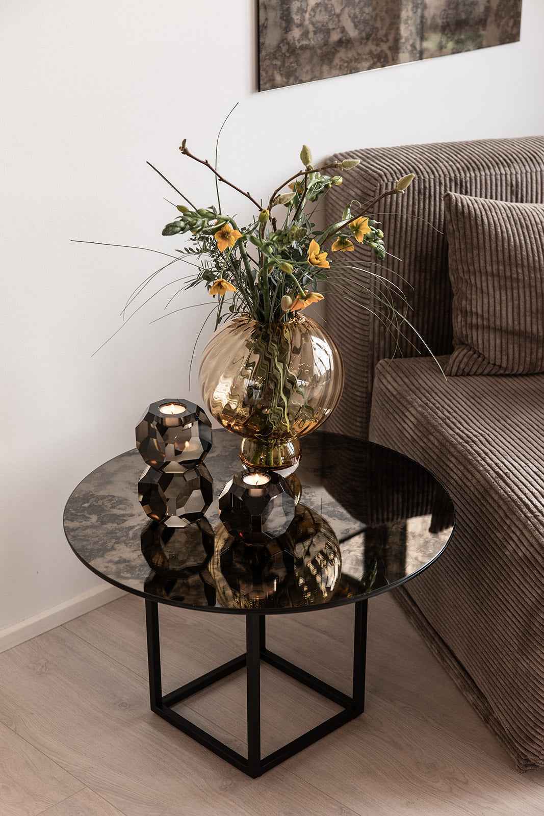 Rundt sofabord med mørk mønstret glasplade og sorte metalben, dekoreret med en ravfarvet glasvase med blomster og to mørke krystal fyrfadsstager, placeret ved siden af en brun fløjlssofa.
