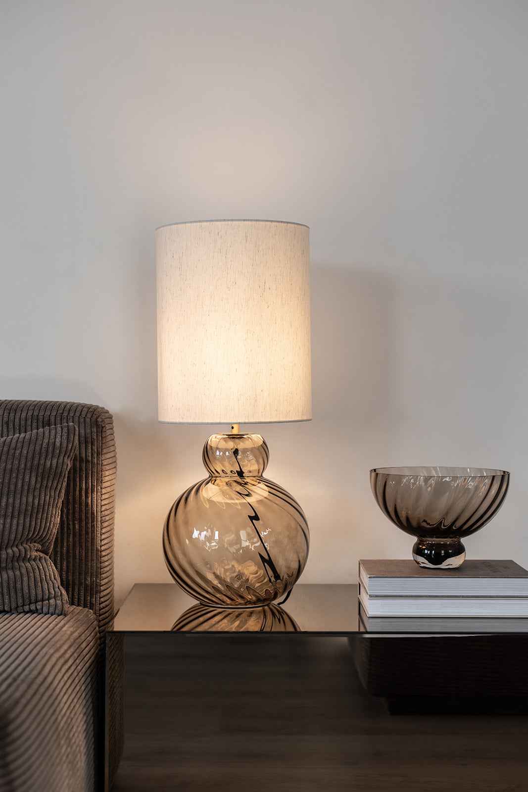 En tændt bordlampe med cylinderformet hvid stofskærm og fod i røgfarvet glas med sorte striber står på et bord ved siden af en sofa med brunriflet stof. På bordet ligger to bøger stablet ovenpå hinanden med en røgfarvet glasskål placeret ovenpå. Baggrunden er en lys væg.